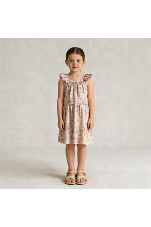 A young girl stands wearing a pastel floral ruffle tiered dress, showcasing a relaxed silhouette and delicate ruffles. The dress is made from organic materials, perfect for sunny days.