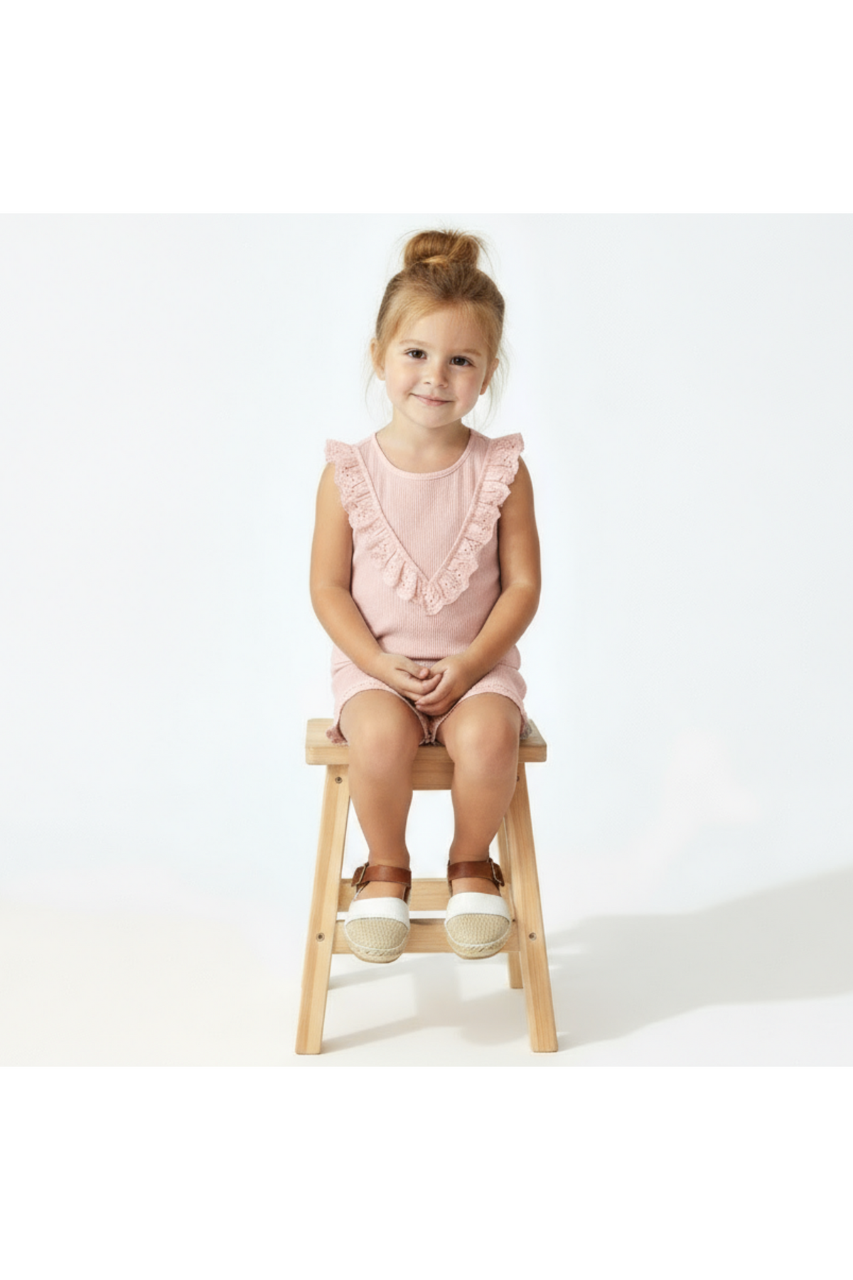 A young girl sits on a wooden stool, wearing a blush organic cotton sleeveless ruffle top.