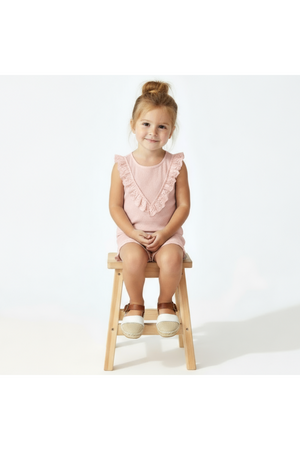 A young girl sits on a wooden stool, wearing a blush organic cotton sleeveless ruffle top.