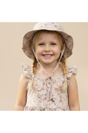 A smiling child wears a pastel floral bucket sun hat made from soft organic cotton. The hat features a wide brim and ties, perfect for sun protection during outdoor activities.