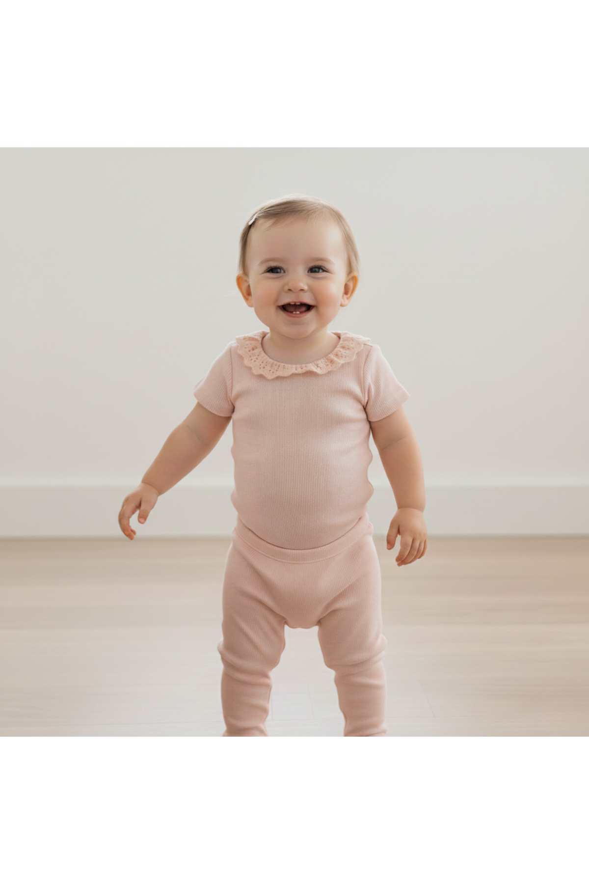 A smiling toddler stands in a soft blush organic cotton onesie with a lace collar, showcasing a cozy ribbed texture. The light, airy setting features a neutral background.