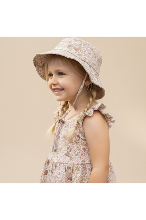 A young child smiles while wearing a pastel floral bucket sun hat made from organic cotton. The hat features a wide brim and is paired with a matching ruffled dress.