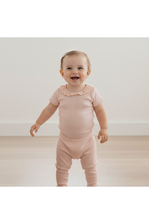 A smiling toddler stands in a soft blush organic cotton onesie with a lace collar, showcasing a cozy ribbed texture. The light, airy setting features a neutral background.