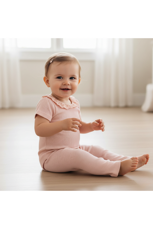 A smiling baby sits on a light wooden floor, wearing a soft blush organic cotton onesie with a lace collar. Sunlight filters through sheer curtains, creating a warm, cozy atmosphere.
