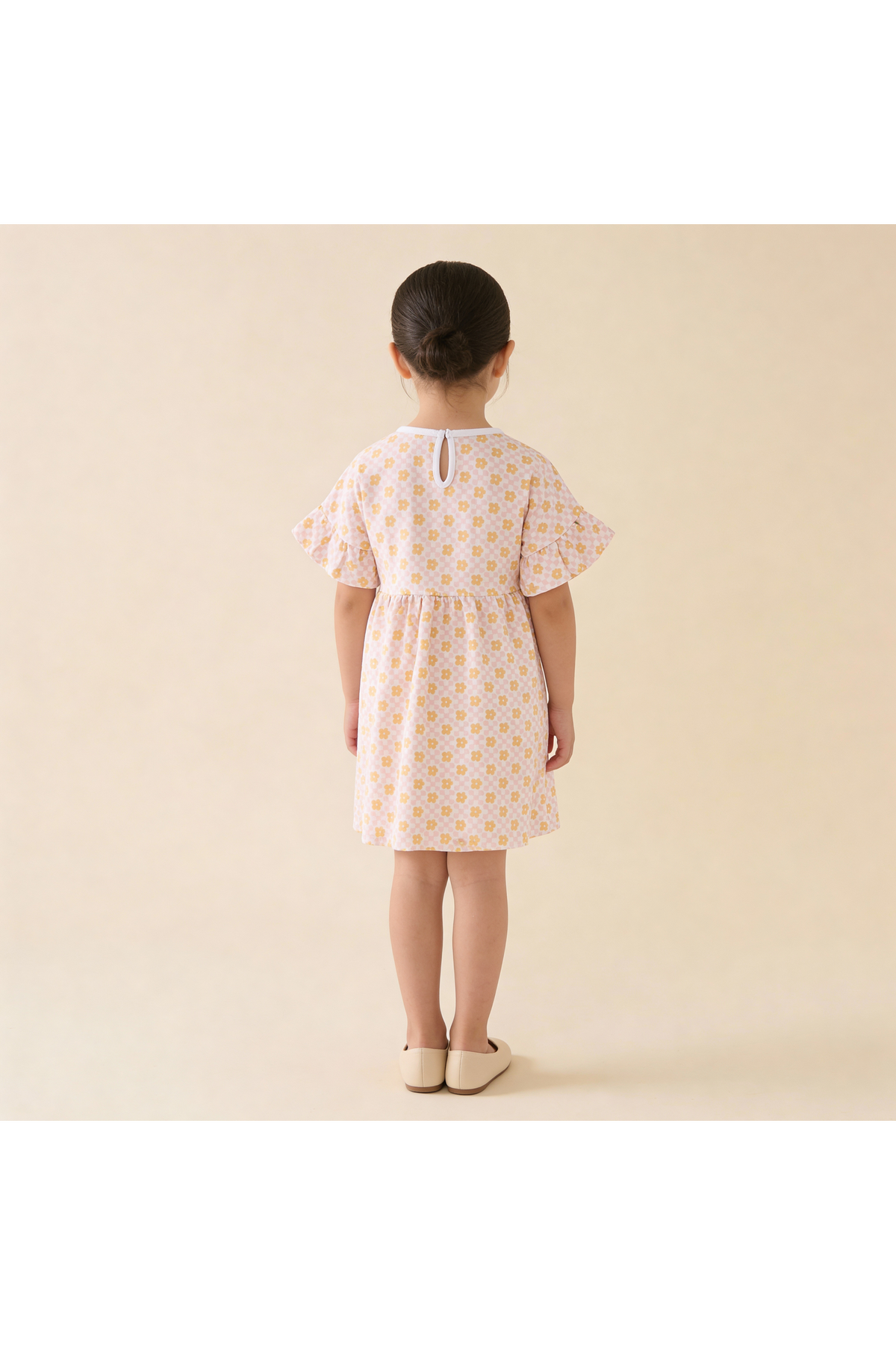 A young girl stands with her back to the camera, wearing a short, organic cotton dress featuring a pop floral print in pastel colors.