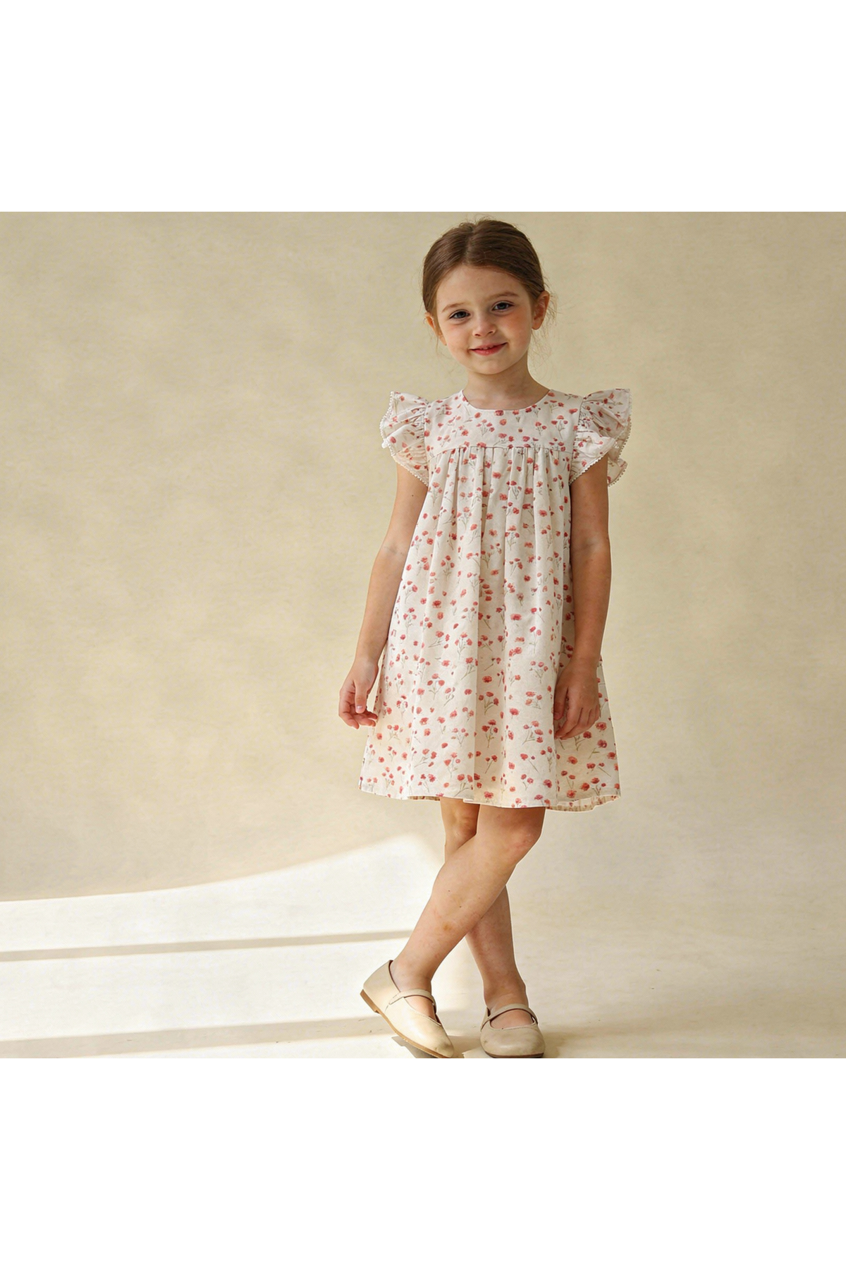 A young girl stands in a light, airy space wearing a sleeveless pink dress adorned with floral patterns.