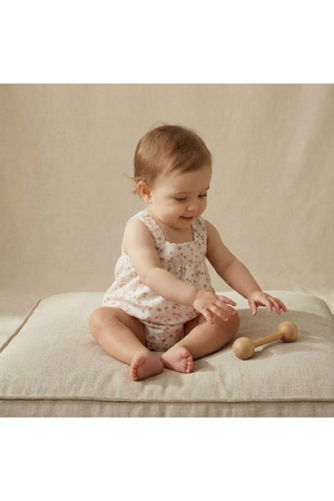 A happy baby sits on a soft, neutral-colored cushion, wearing a floral, sleeveless romper. She plays with a wooden toy, showcasing a relaxed and playful atmosphere.