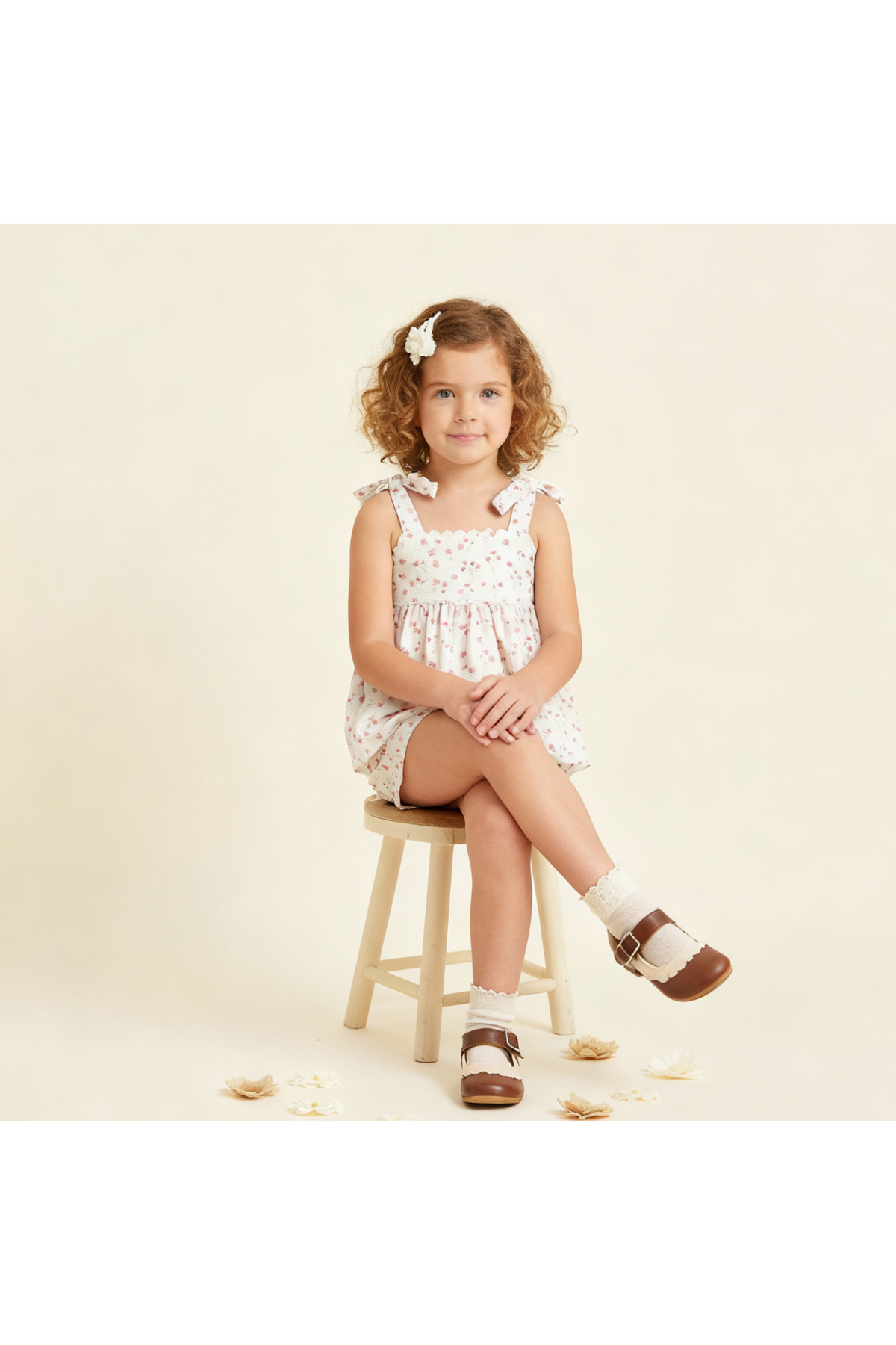 A young girl with curly hair sits on a wooden stool, wearing a floral patterned romper with adjustable straps and brown sandals, showcasing a charming.