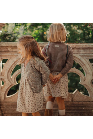Two young girls are outdoors, one wearing a floral print dress and a cozy cardigan, while the other dons a simple gray sweater.