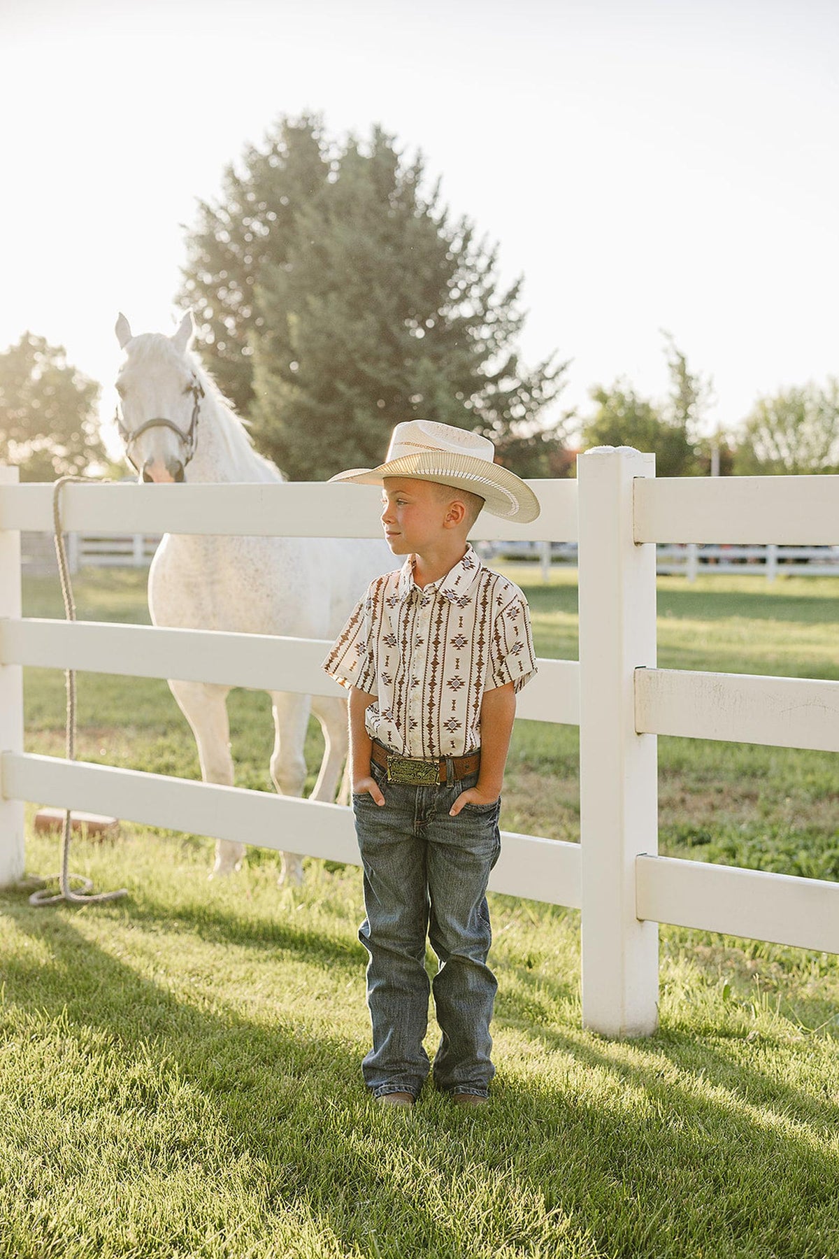 A young boy stands near a white fence, wearing a beige and brown patterned button-up shirt, jeans, and a wide-brimmed hat, with a horse grazing in the background.