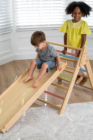 Two children play on a wooden Pikler triangle playset with a slide, featuring brightly colored rungs. The indoor setting has natural light and a soft area rug.