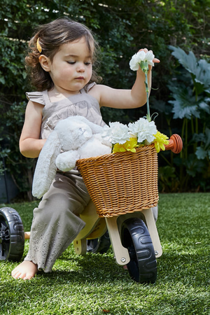 A young girl sits on a wooden balance bike, holding flowers while a stuffed bunny rests in a woven basket. She wears a light-colored outfit and is surrounded by greenery.