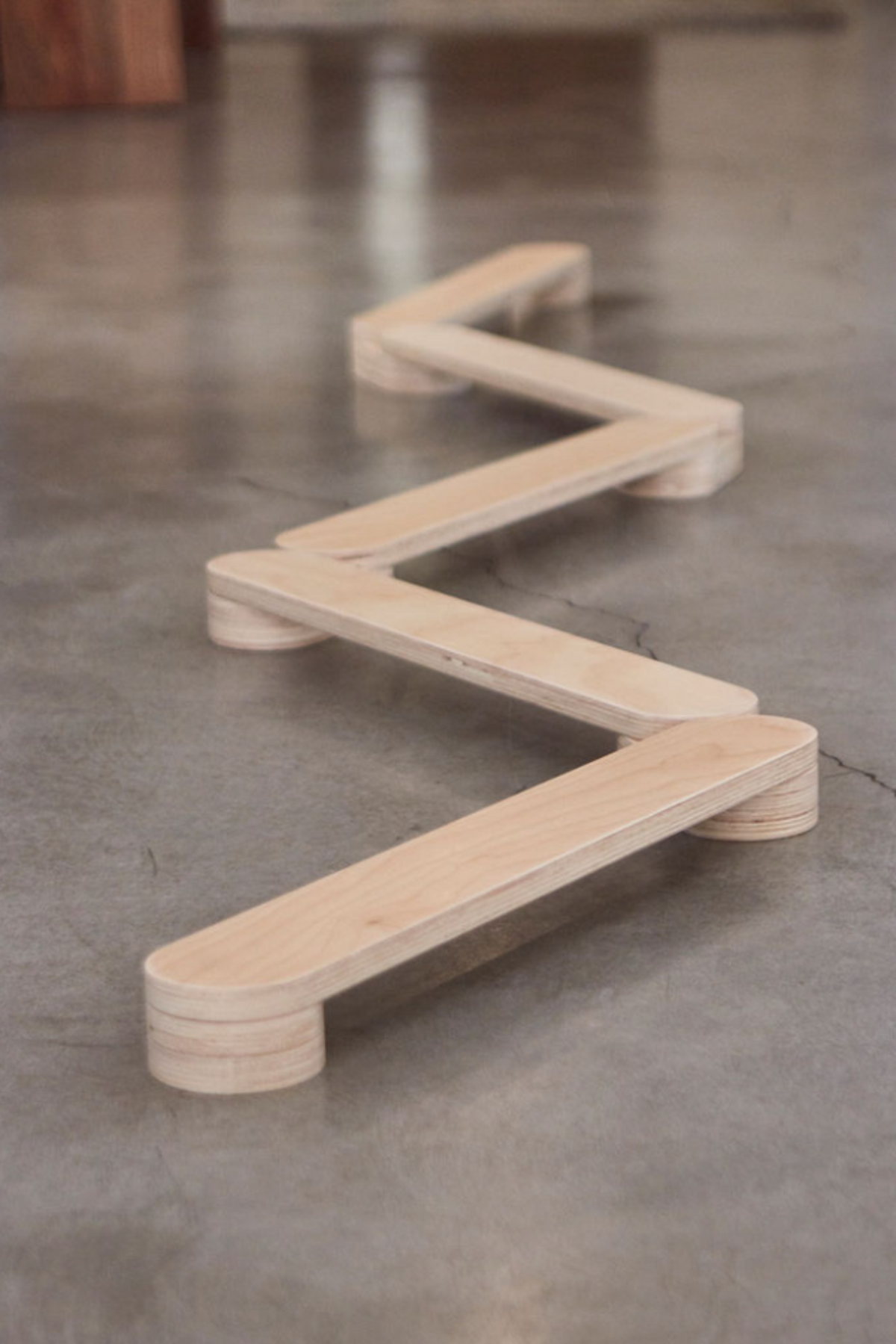 A zigzag wooden balance beam with a smooth finish rests on a concrete floor, showcasing its natural wood grain. This sustainable play structure promotes coordination and active movement for children.
