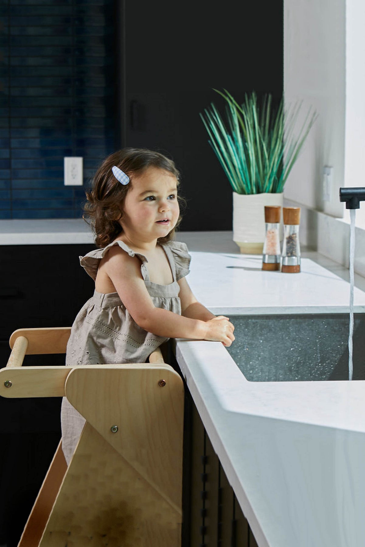 A young girl stands on a wooden educational tower by a kitchen sink, smiling as she engages with water.