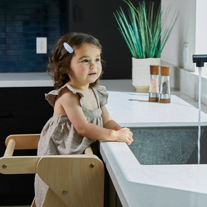 A young child stands at a kitchen sink, smiling while using a wooden foldable educational tower. The modern kitchen features dark cabinetry and a vibrant green plant in the background.