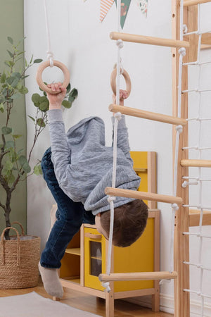 A child in a gray hoodie hangs upside down from wooden rings attached to a natural wood jungle gym in a bright indoor play space, surrounded by plants.