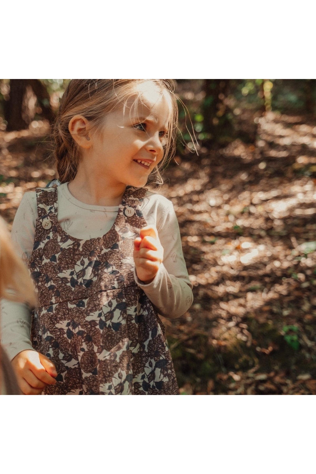 A young girl smiles while wearing a floral corduroy dress with adjustable straps, surrounded by a natural, wooded setting. Sunlight filters through the trees, enhancing the cheerful atmosphere.