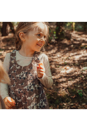A young girl smiles while wearing a floral corduroy dress with adjustable straps, surrounded by a natural, wooded setting. Sunlight filters through the trees, enhancing the cheerful atmosphere.