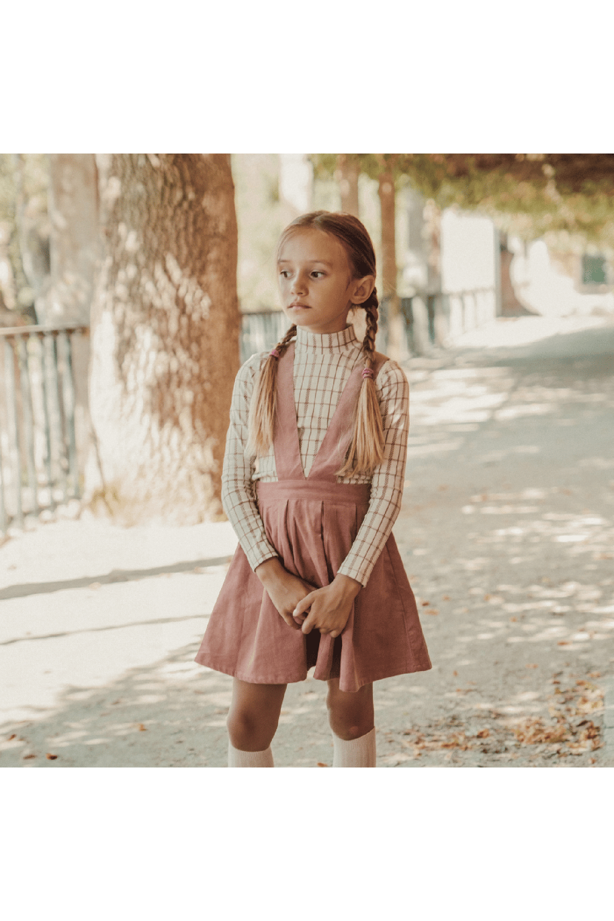 A child stands outdoors in a soft, striped turtleneck and a mauve dress, surrounded by trees. The outfit is playful and suitable for various casual occasions.