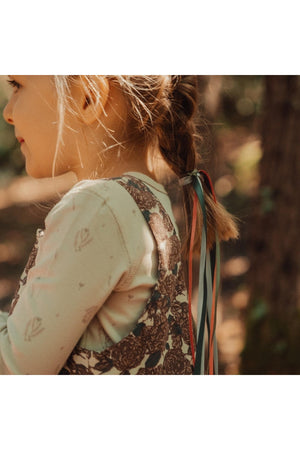A young child with braided hair wears a floral corduroy dress, showcasing adjustable straps in earthy colors. The setting features a natural, wooded background, enhancing the dresss charming appeal.