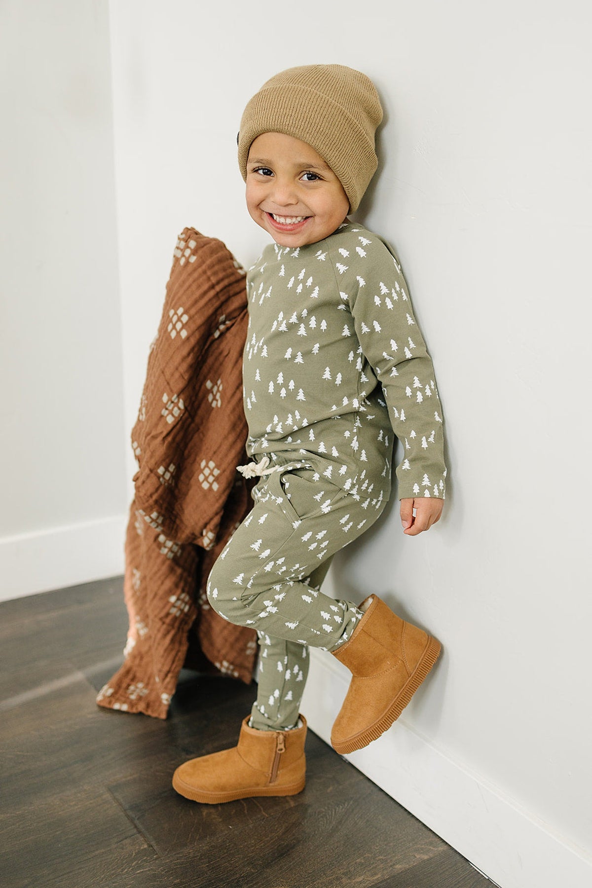 Smiling child in green pajamas with white tree print, wearing a tan beanie and brown boots, holding a quilt.