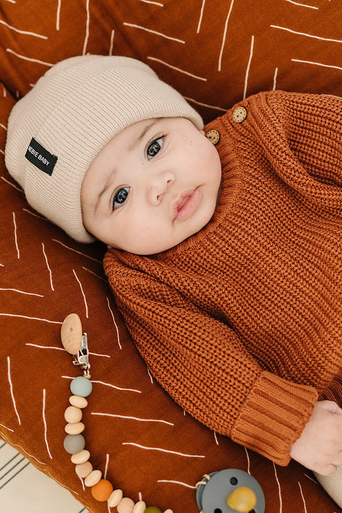 Baby wearing a beige knit hat and rust sweater, lying on a rust-colored blanket with a wooden bead pacifier clip.