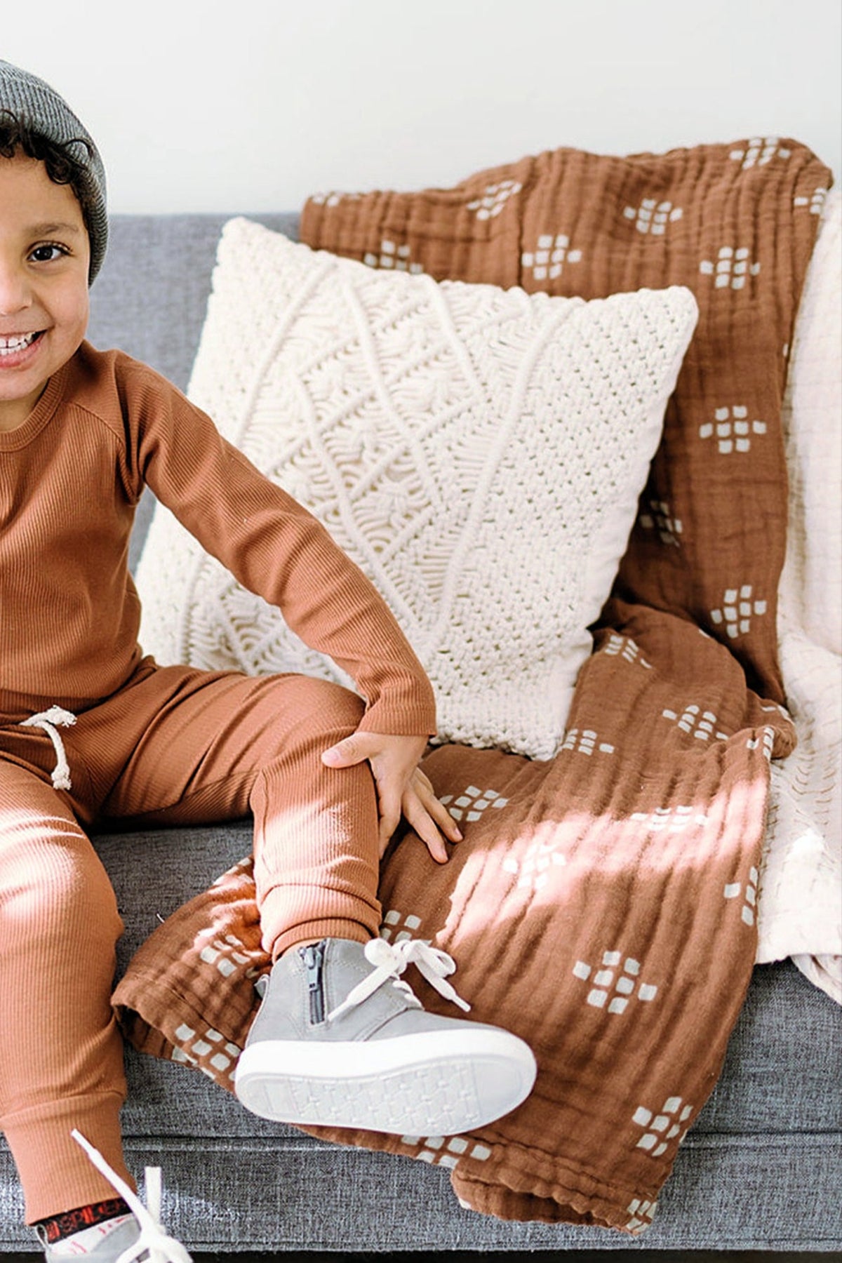 A smiling child in brown pajamas sits on a gray sofa with a textured cream pillow and patterned brown quilt.