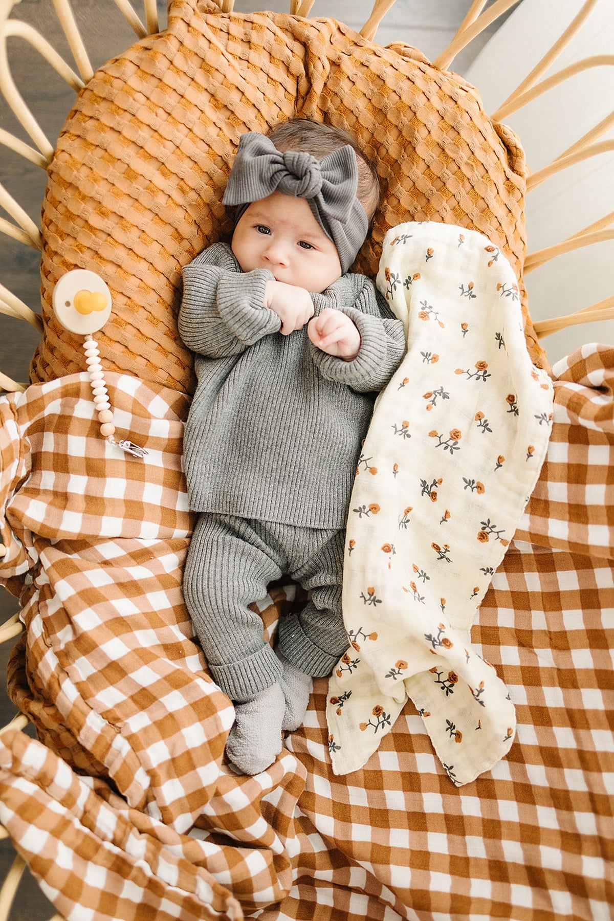 A baby in a gray outfit and headband lies on a rust and white gingham quilt.