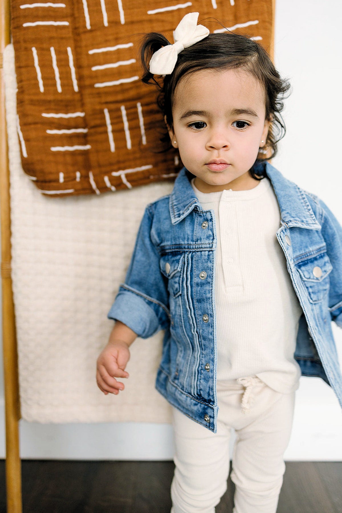 A toddler wearing a blue denim jacket and white outfit stands in front of a brown and white blanket.