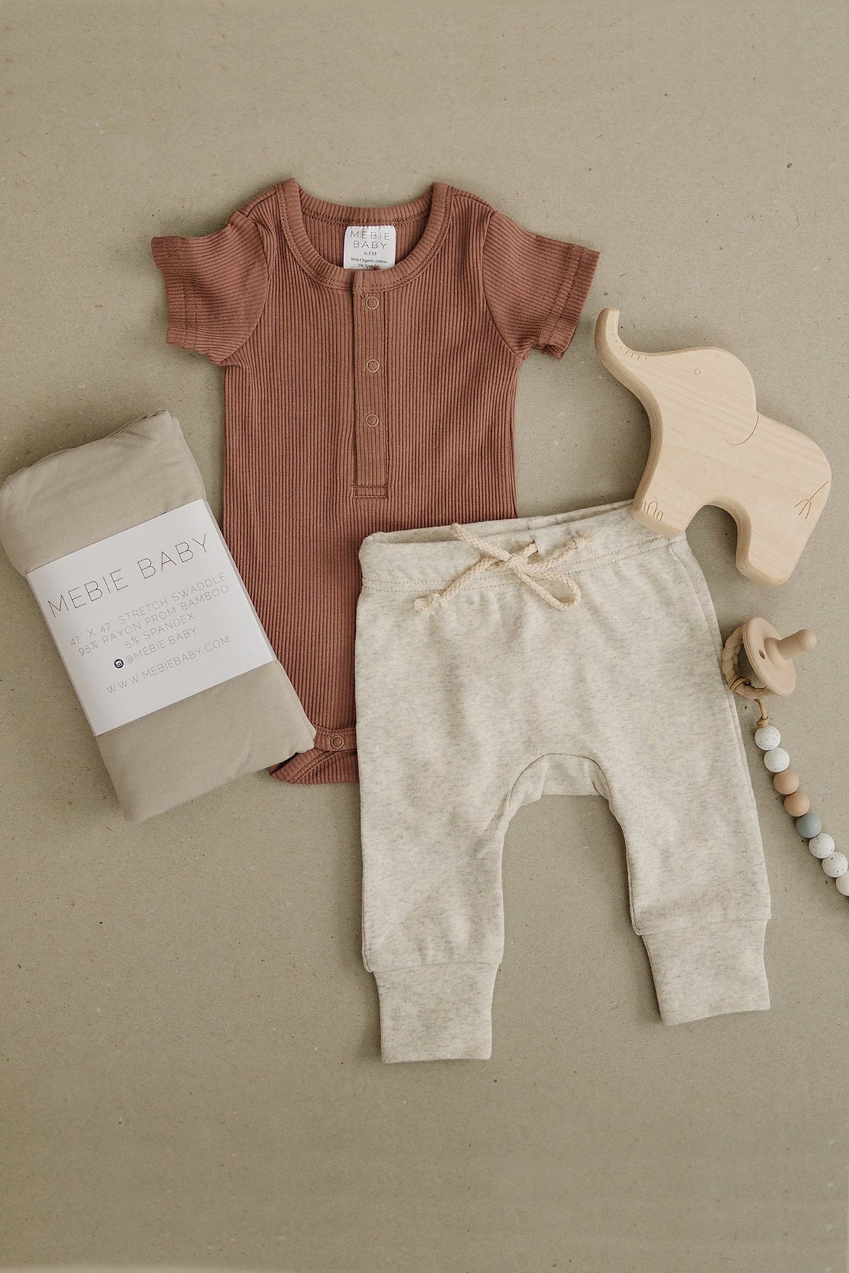 Dusty rose ribbed baby bodysuit with snaps, beige pants, wooden elephant toy, and a Mebie Baby swaddle.