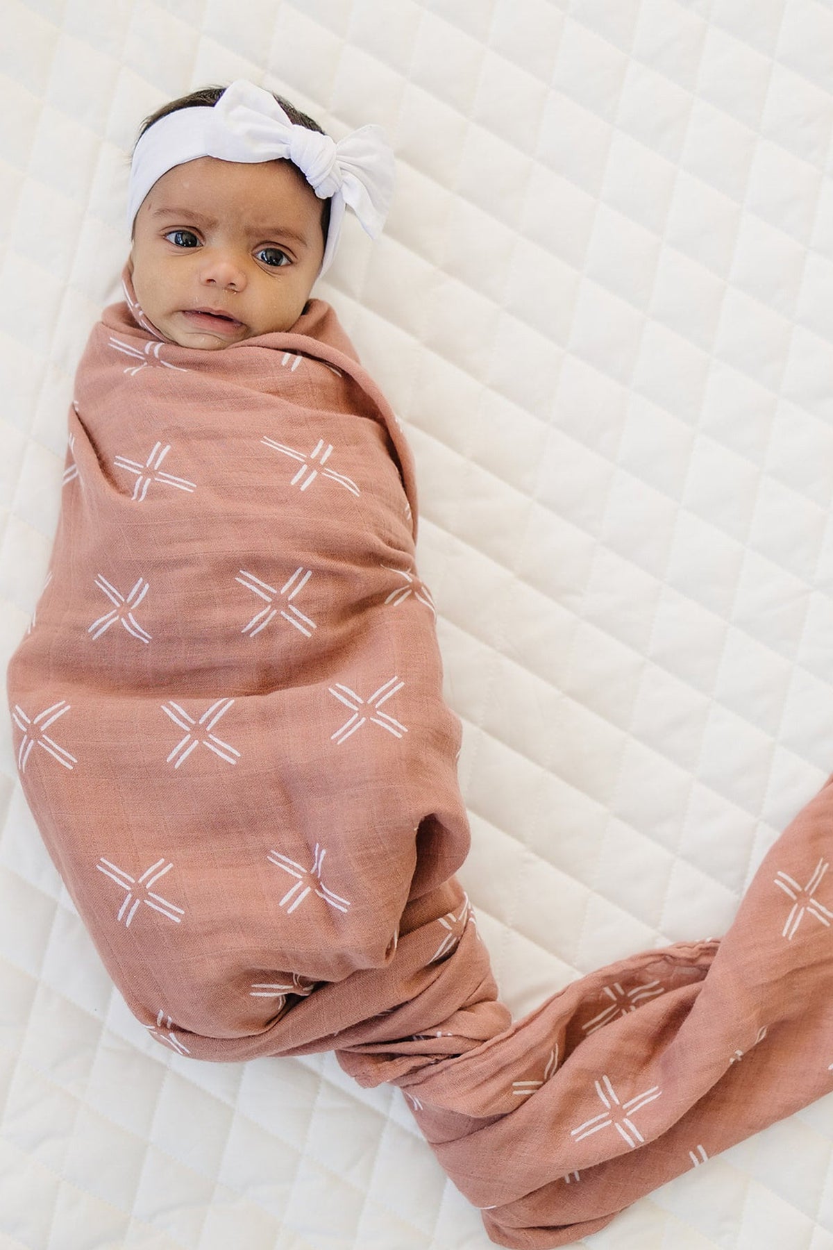 A baby wrapped in a peach-colored muslin swaddle with white patterns, lying on a quilted white surface.