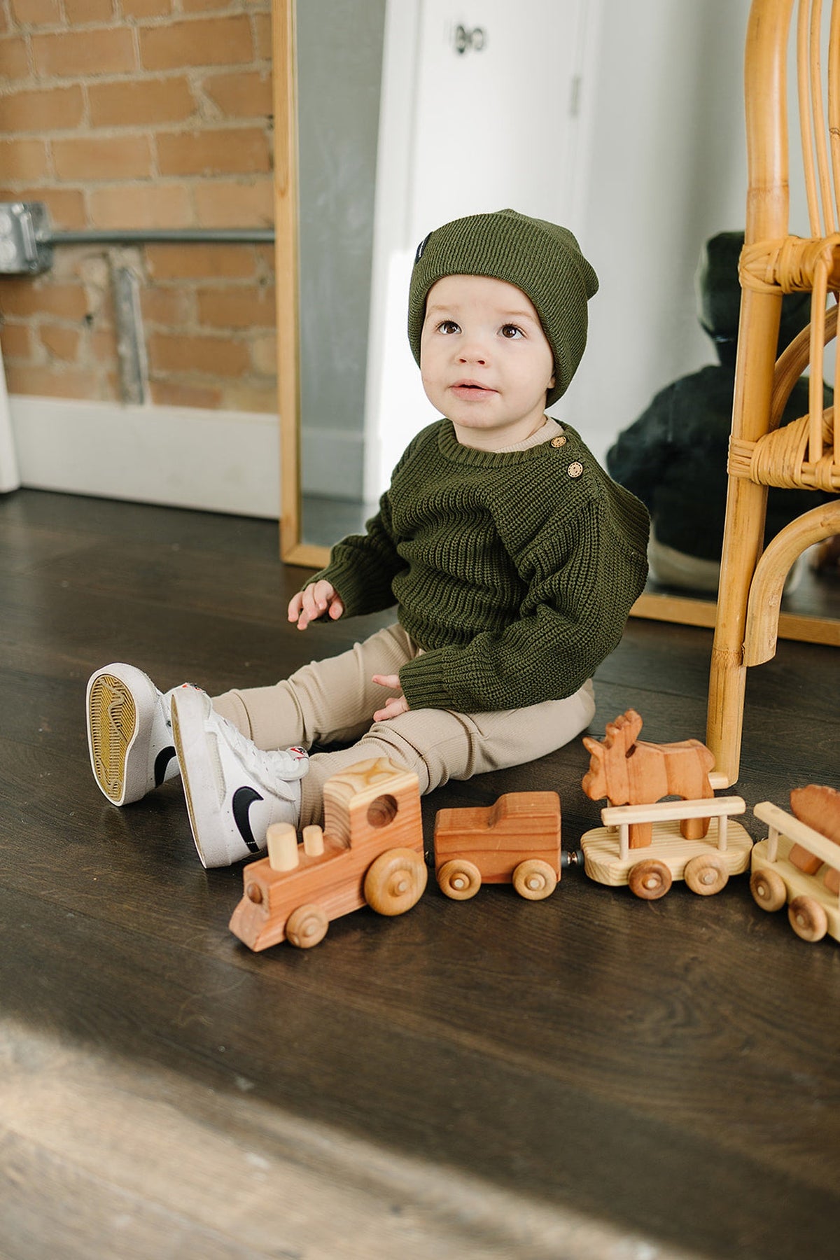 A toddler in oatmeal leggings and a green sweater sits on a wooden floor with wooden toy trains.