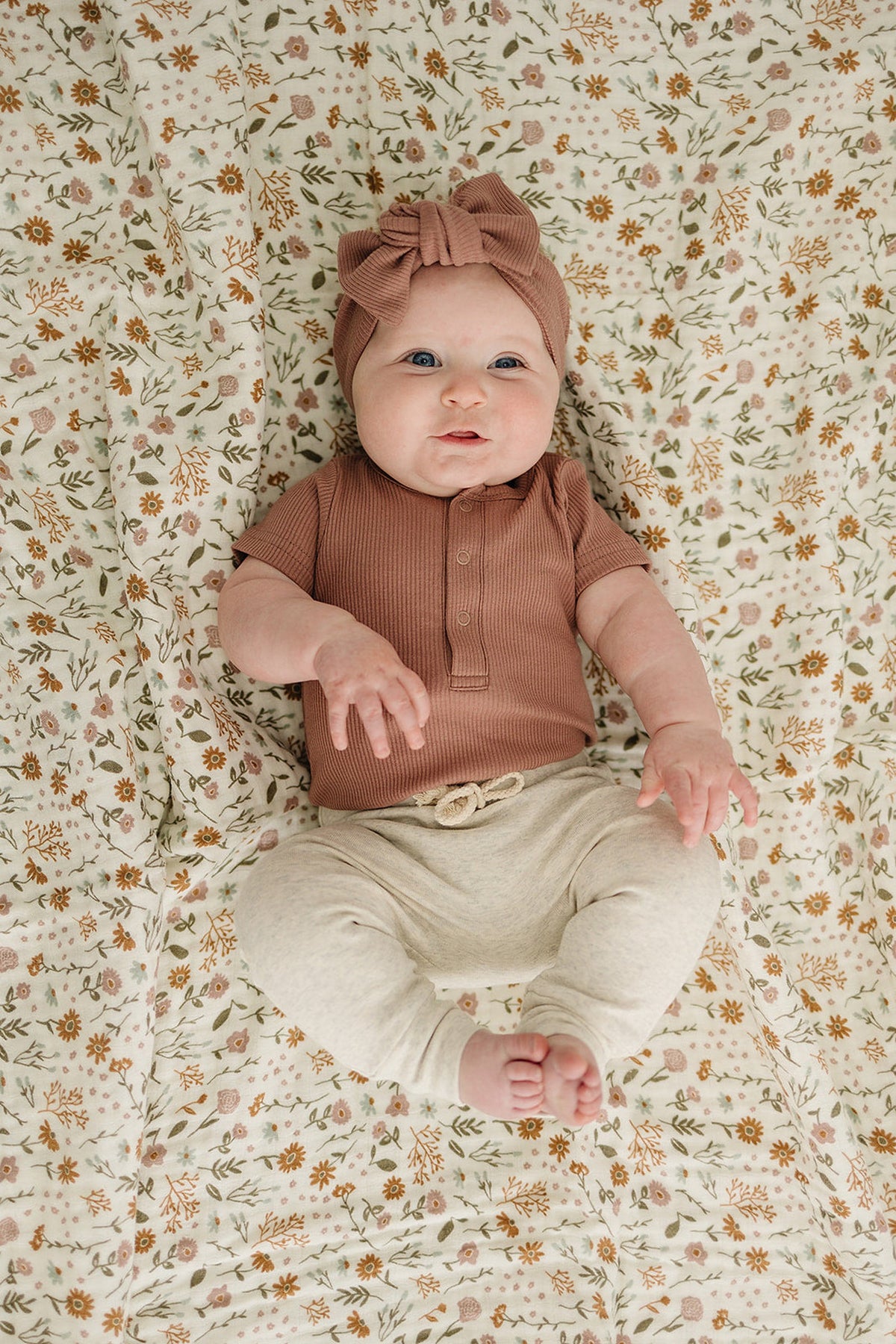 A baby wearing a dusty rose ribbed bodysuit and headband lies on a floral-patterned blanket, smiling.