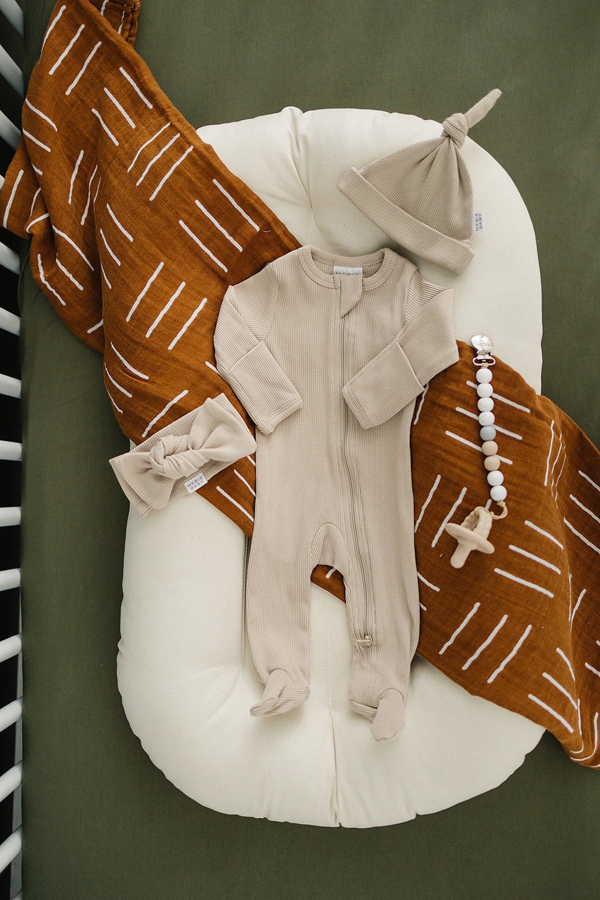 Beige baby outfit with matching hat and bow, laid on a white cushion with a rust-colored blanket.