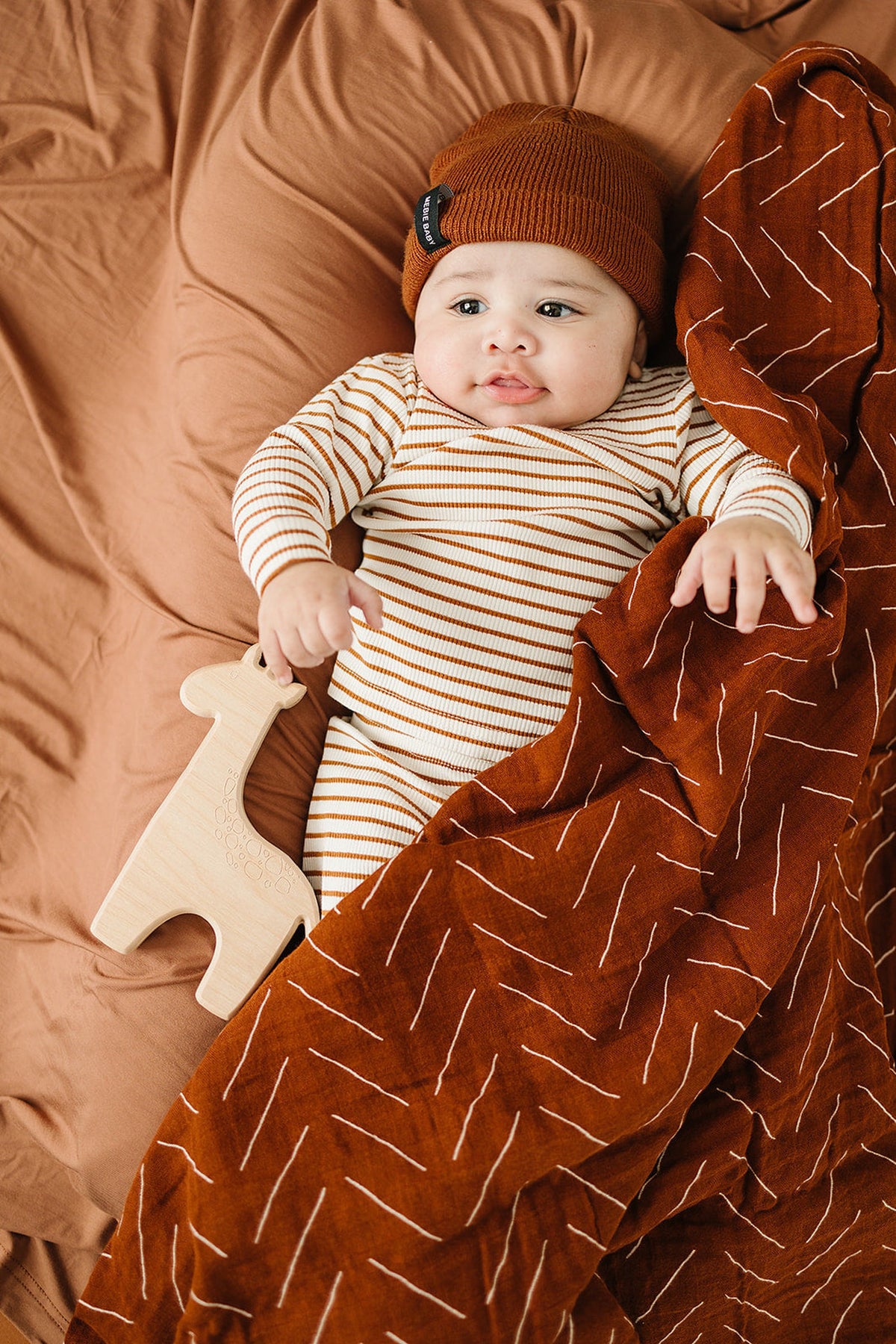 A baby in a striped onesie and rust beanie lies on a rust muslin blanket, holding a wooden giraffe.