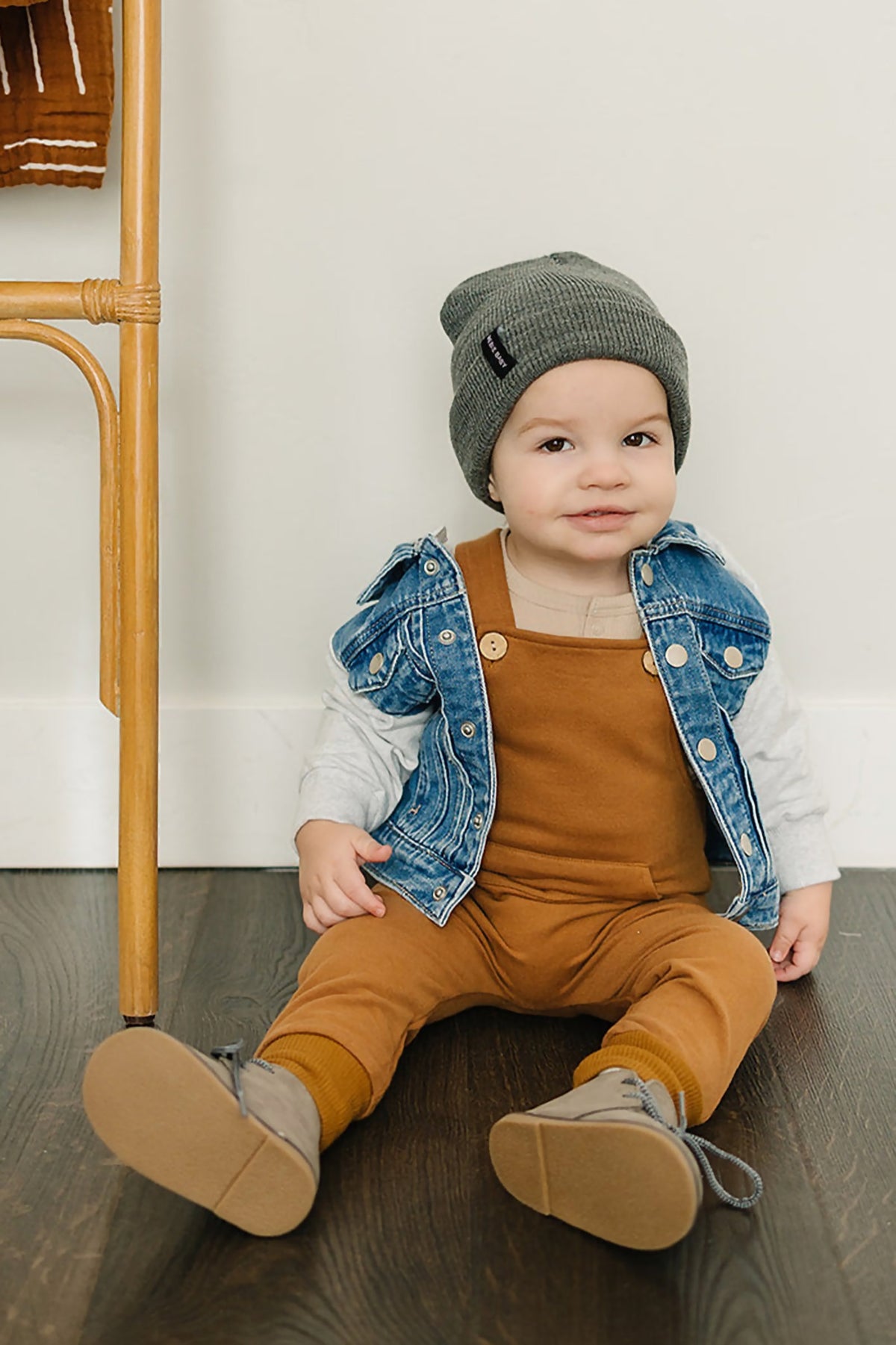 A toddler wearing a denim vest over a brown outfit and gray beanie sits on a wooden floor smiling.
