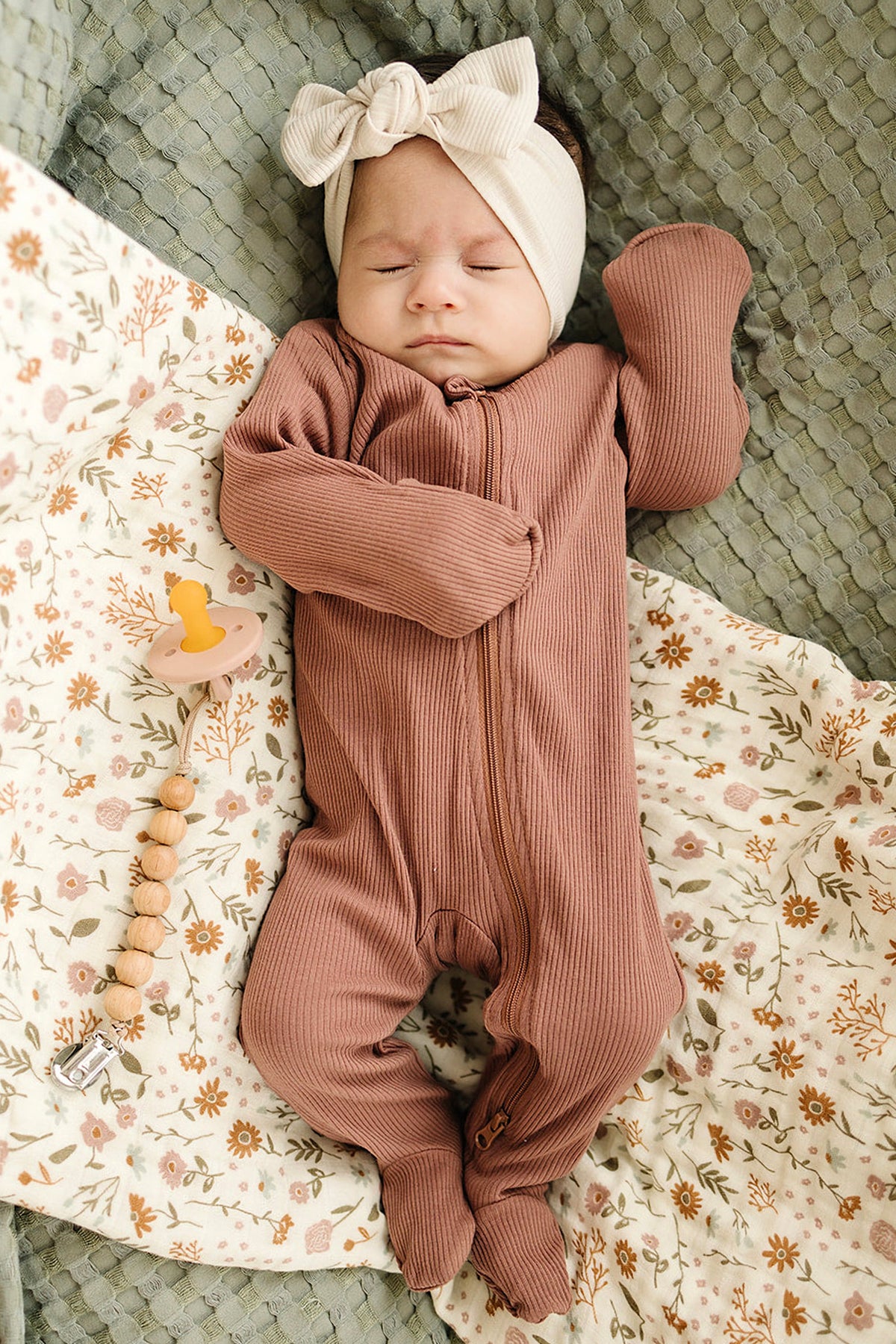 A baby wearing a dusty rose ribbed zippered pajama and cream headband, lying on floral and textured blankets.