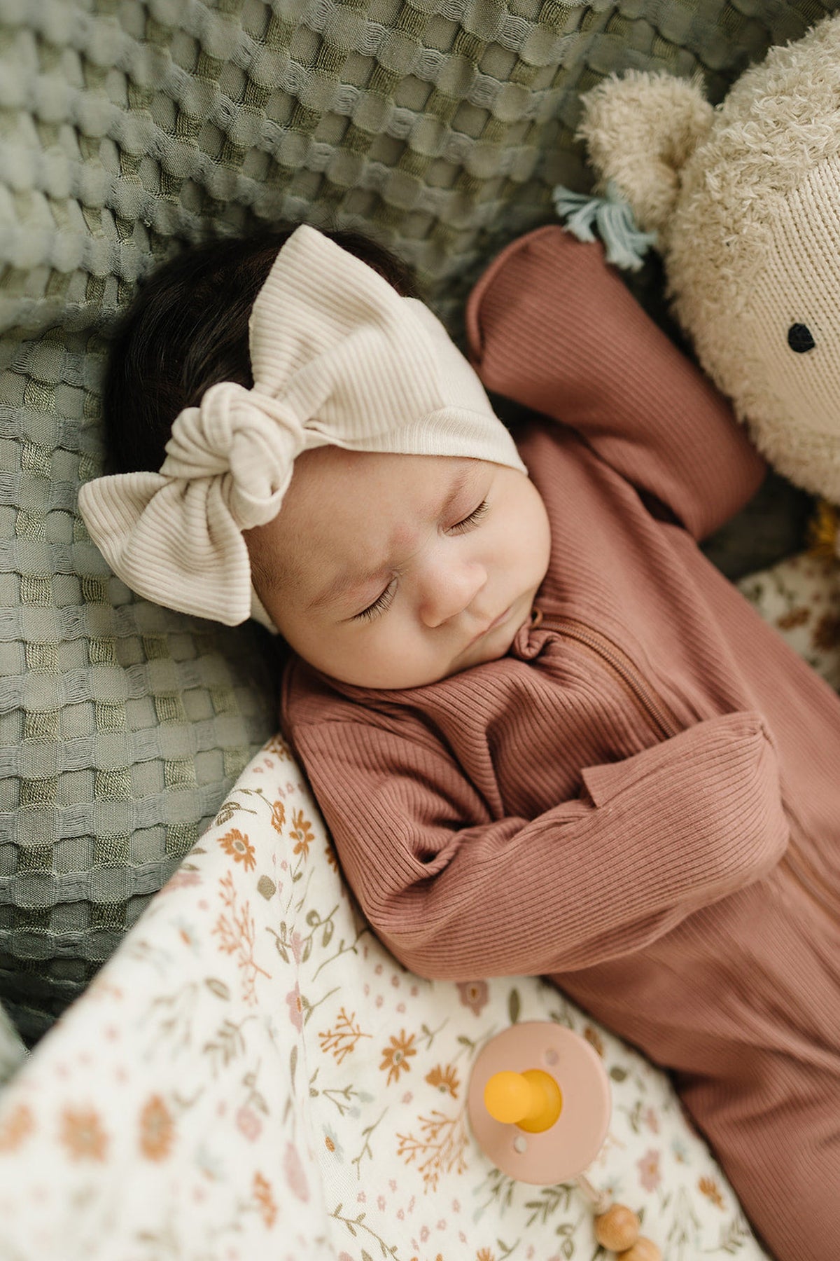 A baby wearing a vanilla ribbed head wrap and rust-colored outfit sleeps beside a plush toy and pacifier.