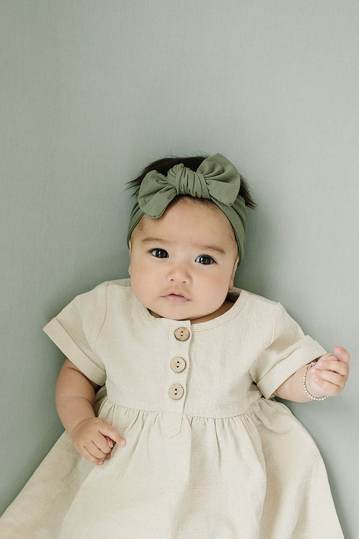 Baby wearing an olive head wrap and cream dress with wooden buttons, lying on a soft green background.