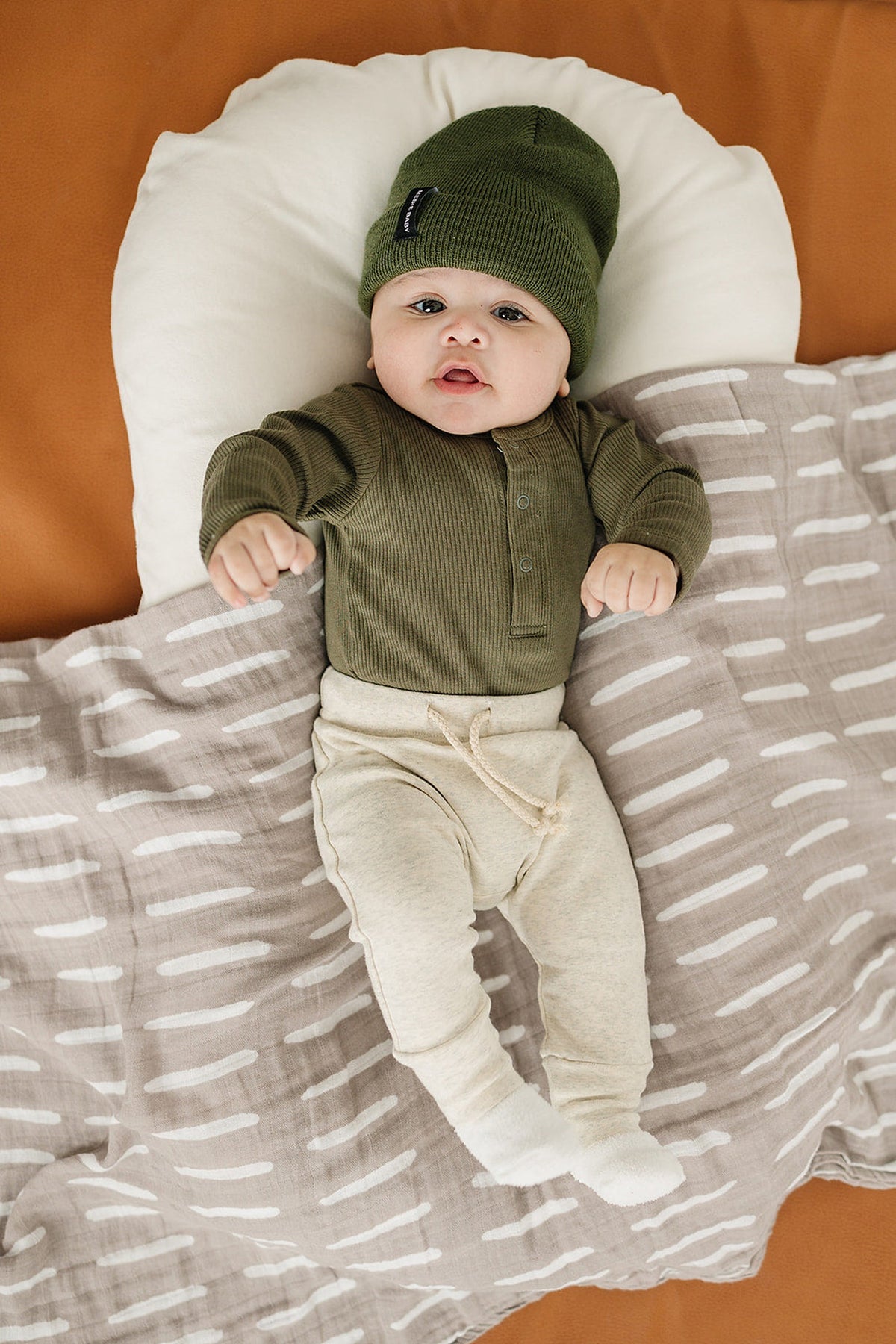 Baby in a green ribbed long-sleeve bodysuit and hat, lying on a patterned blanket with a neutral background.