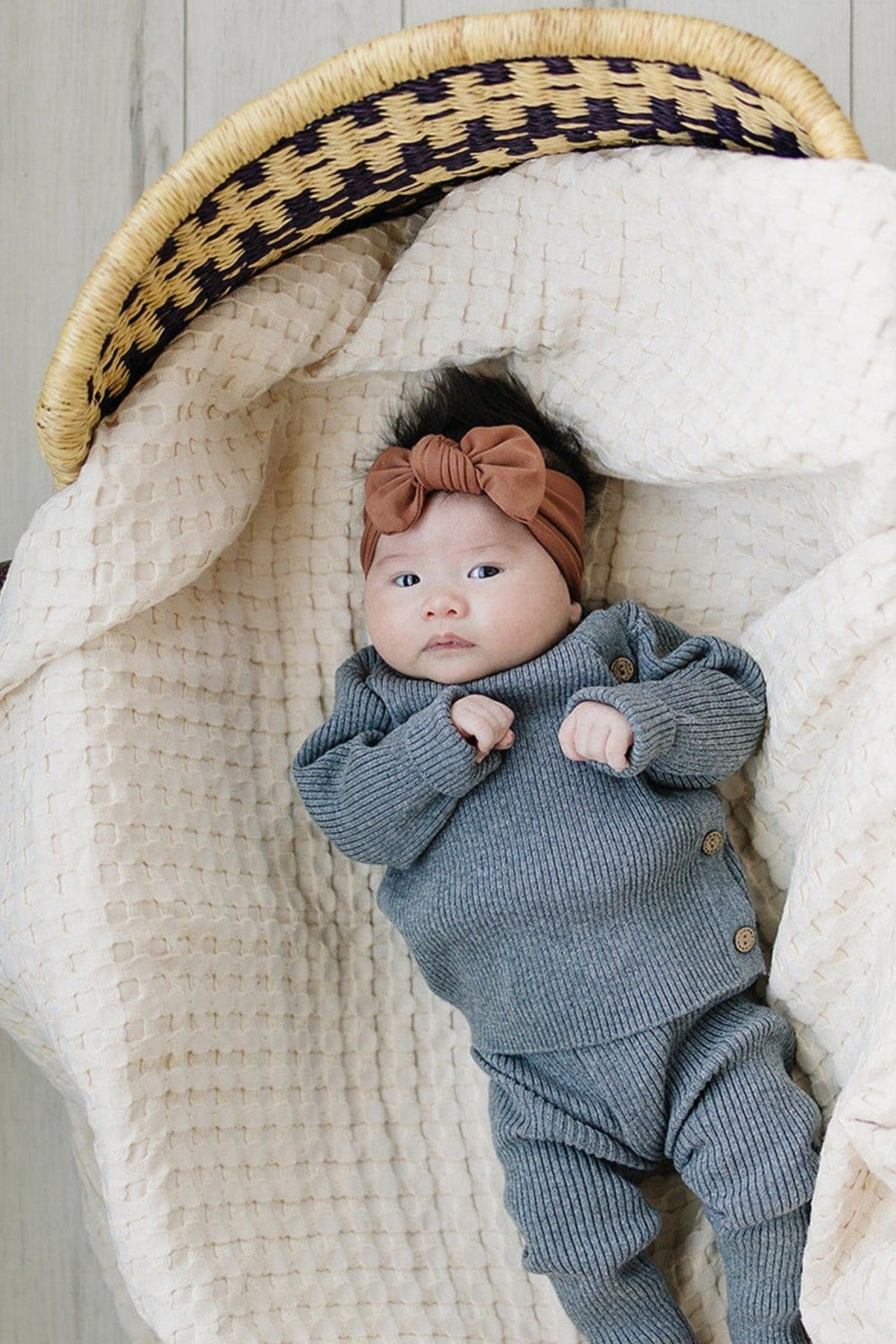 A baby wearing a rust-colored head wrap and gray outfit lies in a woven basket with a textured blanket.