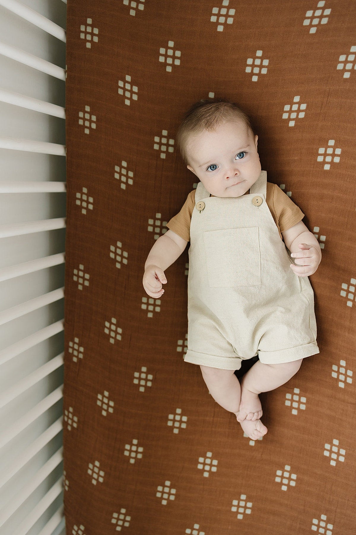 A baby lies on a brown muslin crib sheet with white geometric patterns, wearing a beige romper.