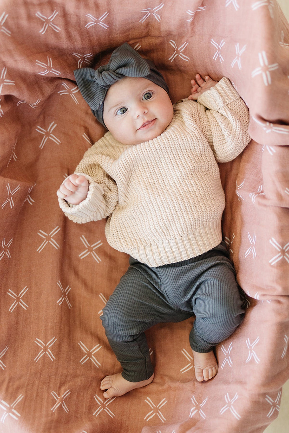 Baby in a beige sweater and charcoal leggings lies on a patterned pink blanket, wearing a matching headband.