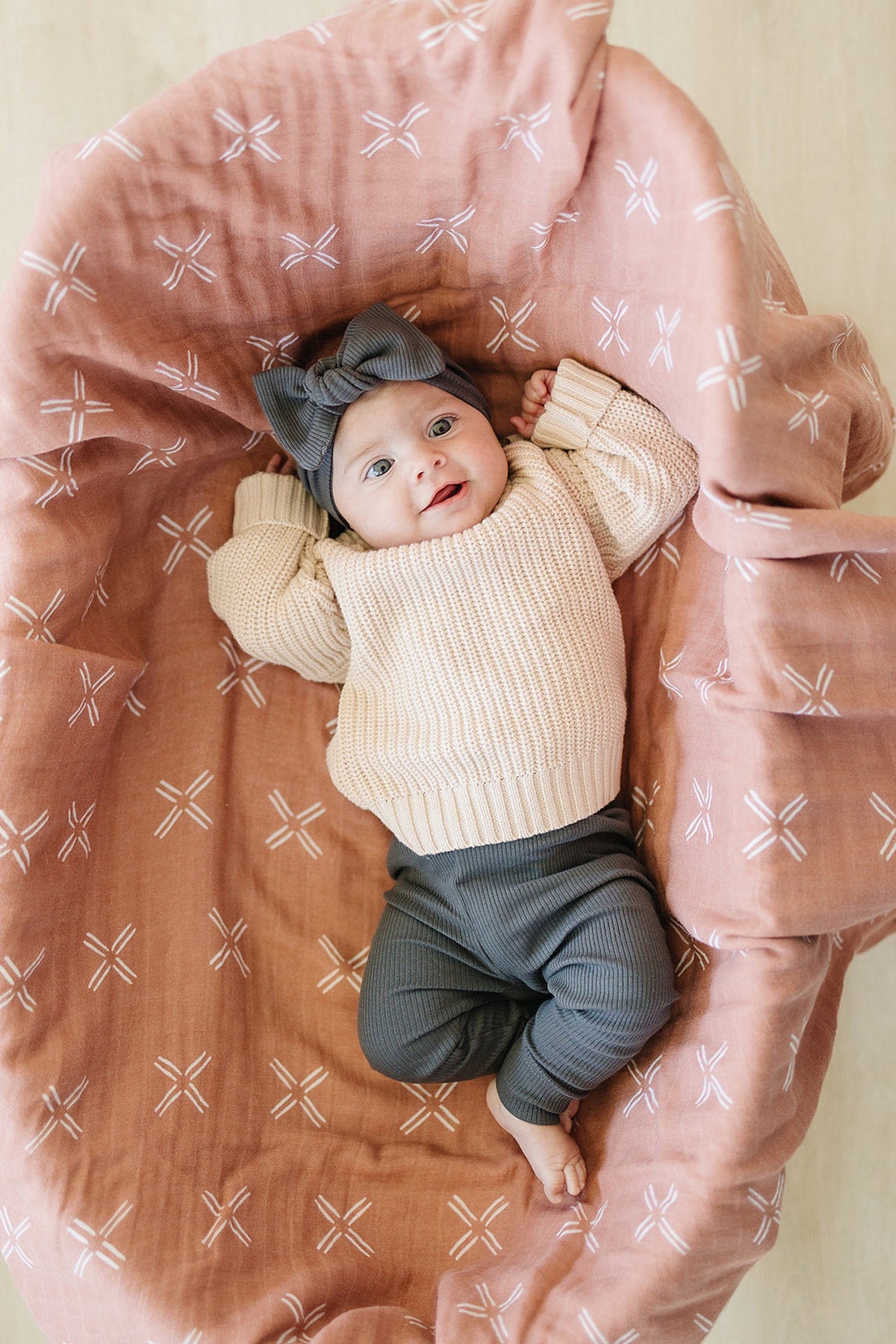 A baby wearing charcoal leggings and a cream sweater lies on a patterned pink blanket, smiling.