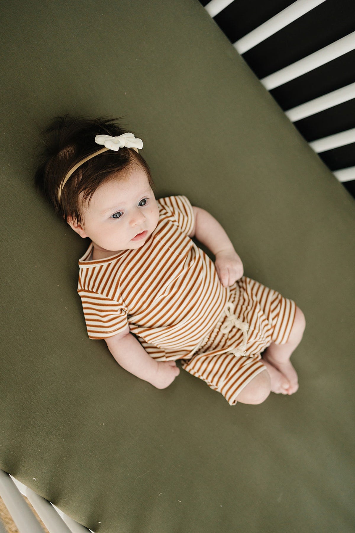 Baby in a striped outfit and bow headband lying on an olive crib sheet.