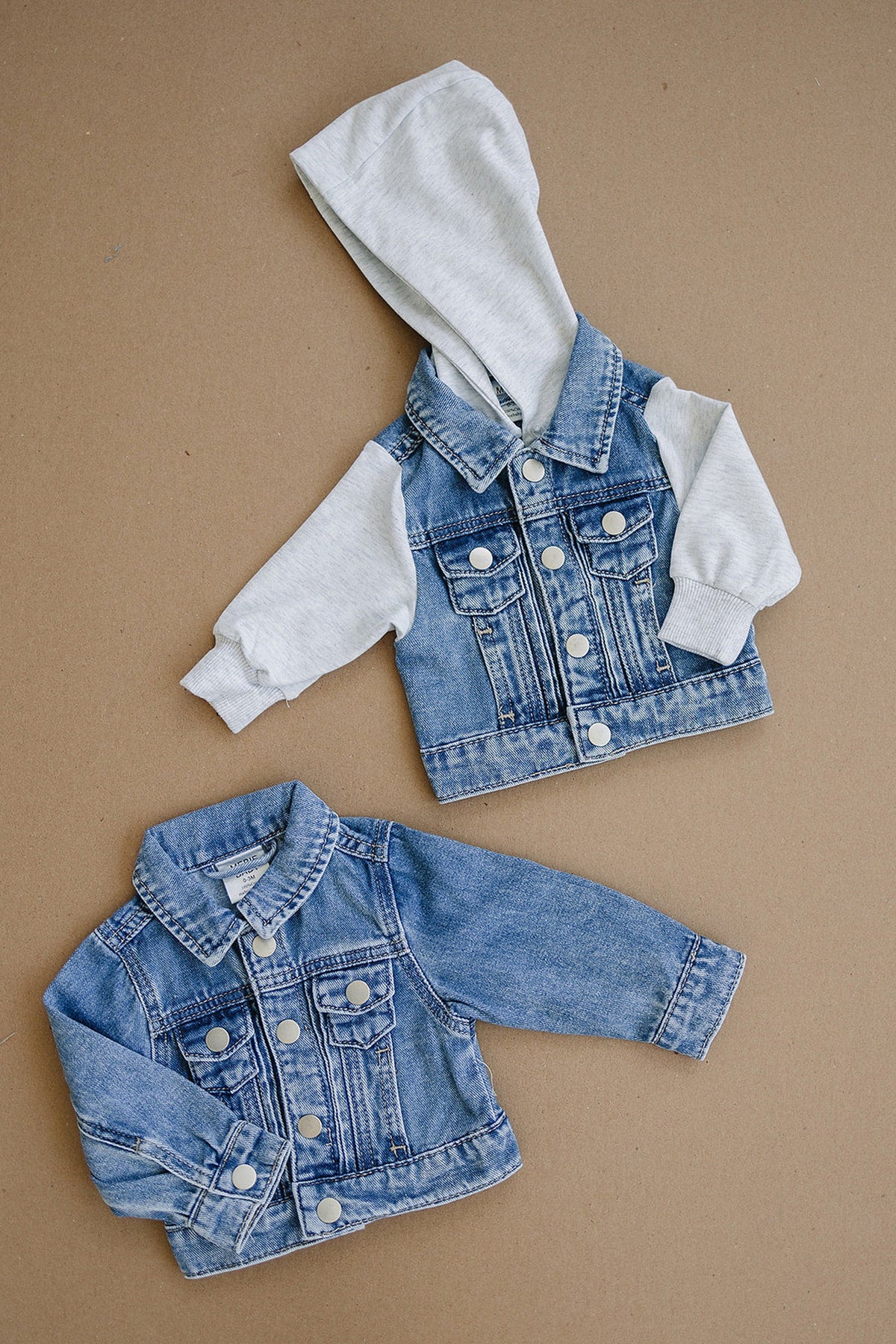 Two baby denim jackets on a brown surface; one with a grey hoodie, the other classic denim style.