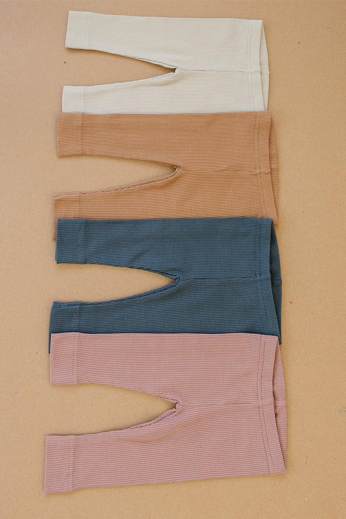 Four pairs of ribbed baby leggings in cream, tan, blue, and dusty rose, displayed on a beige background.