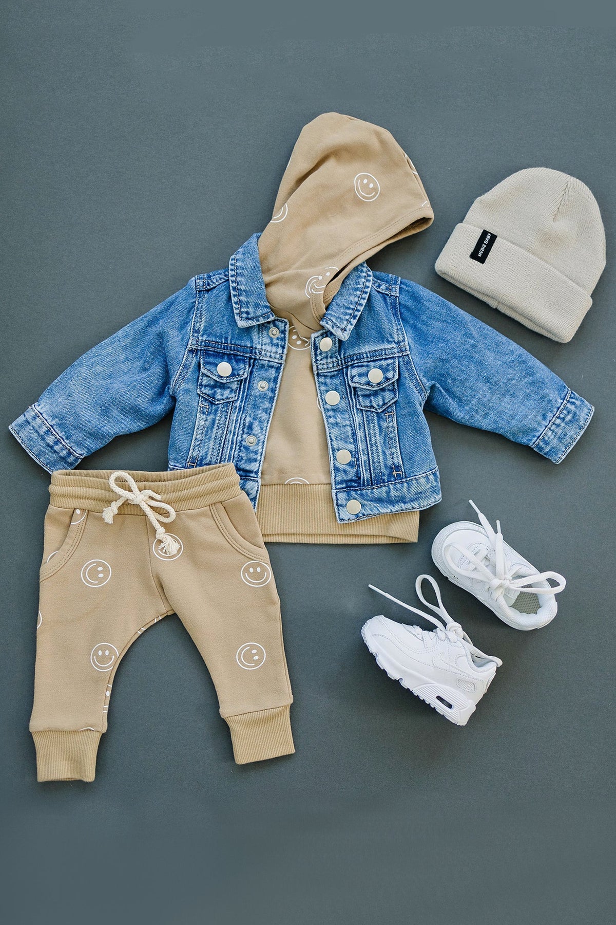 Blue denim jacket, tan hoodie with smiley faces, matching joggers, cream beanie, and white sneakers on gray background.