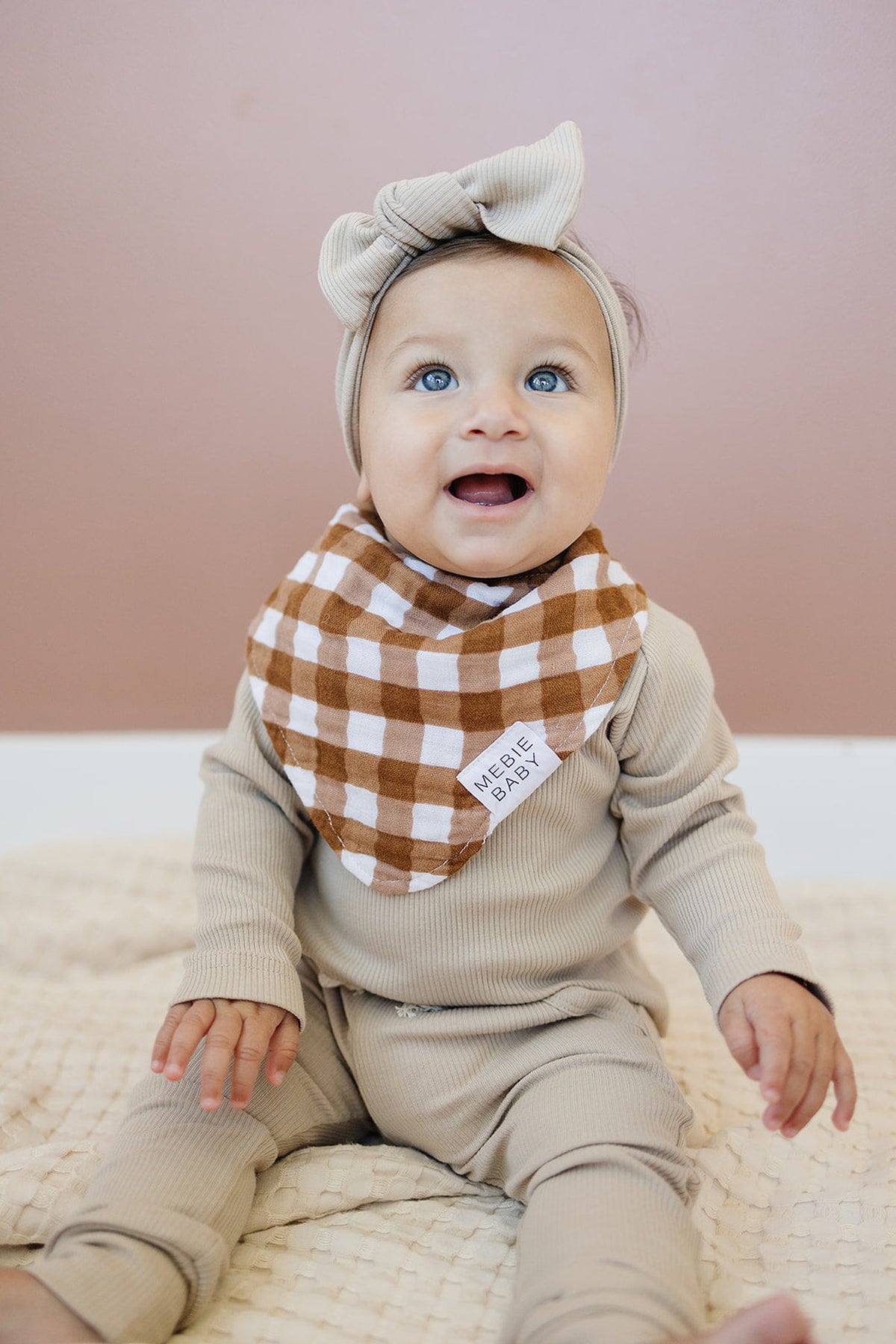 A baby wearing a beige outfit and headband with a brown gingham Mebie Baby bib, sitting happily indoors.