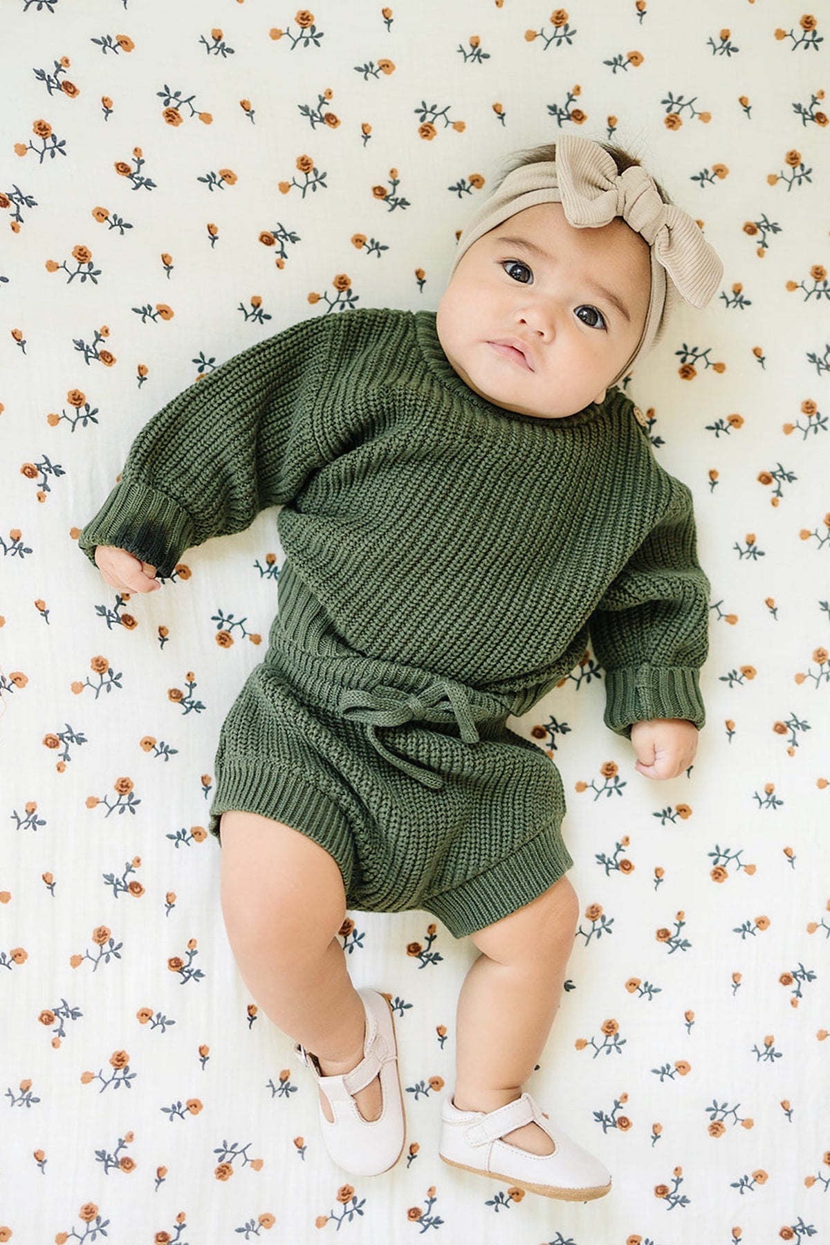 Baby wearing a green knit outfit and beige bow headband lies on a floral-patterned blanket.
